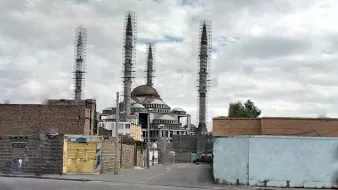 L'image montre un grand bâtiment au centre, semblable à une mosquée, avec plusieurs minarets élancés qui s'élèvent vers le ciel. Le bâtiment a un dôme proéminent et semble être en construction ou en rénovation, avec des échafaudages visibles. En arrière-plan, le ciel est nuageux, ce qui donne à la scène une atmosphère un peu sombre. À l'avant, on peut apercevoir des structures et des murs en briques, ainsi qu'une route. L'ensemble de la composition évoque un mélange d'architecture traditionnelle et moderne.