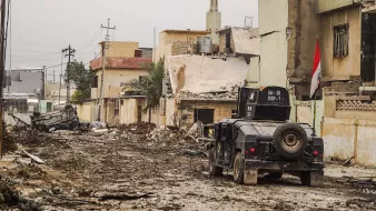 L'image montre une rue dévastée, probablement dans une zone de conflit. Au premier plan, on voit un véhicule militaire sur une route boueuse, entouré de débris. Les bâtiments en arrière-plan semblent endommagés, avec des murs effondrés et des structures incomplètes. Une mosquée est visible, avec son minaret se dessinant contre le ciel. Un drapeau irakien est aussi présent, indiquant que la scène se déroule en Irak. L'atmosphère générale semble sombre et dévastée, reflétant les conséquences d'un conflit armé.