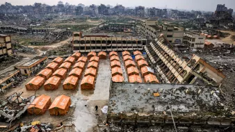 L'image montre un paysage de destruction, avec des bâtiments en ruines en arrière-plan. Au premier plan, des tentes orange sont disposées en rangées sur un terrain dévasté, témoignant d'une situation de crise ou de catastrophe.