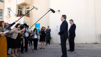 Nell'immagine si può vedere una scena di una conferenza stampa all'aperto. Due persone, vestite in abiti eleganti, sono in piedi di fronte a un muro chiaro, mentre un gruppo di giornalisti le circonda. Alcuni giornalisti stanno utilizzando microfoni e telecamere per registrare o fare domande. Dietro di loro, ci sono alcuni cartelli e avvisi su un edificio. L'atmosfera sembra formale e professionale, con un focus sulle dichiarazioni dei due individui in primo piano.