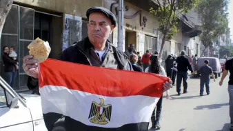 L'image montre un homme tenant un drapeau égyptien, avec le symbole de l'Égypte au centre. Il tient également un morceau de pain dans l'autre main. Demain la ville est animée, avec plusieurs personnes en arrière-plan, ce qui suggère une atmosphère de rassemblement ou de manifestation. L'homme semble sérieux et déterminé, exprimant un fort sentiment national. L'environnement urbain, avec des bâtiments et des passants, reflète la vie quotidienne en Égypte.