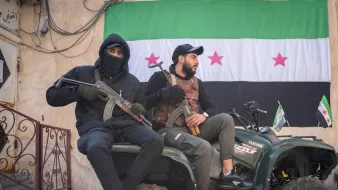 Deux hommes assis sur un quad, l'un tenant une arme, l'autre un drapeau. En arrière-plan, un mur avec un grand drapeau portant des étoiles rouges sur fond vert et noir. L'ambiance semble militaire et tendue.