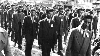 L'image montre un groupe d'hommes marchant ensemble dans une rue pavée. Tous portent des costumes et semblent sérieux. Ils avancent en rang, avec des badges sur leur veste. En arrière-plan, on peut distinguer quelques bâtiments et enseignes, ce qui donne un sens d'époque à la scène. L'atmosphère peut paraître solennelle, suggérant possiblement une manifestation ou un événement organisé.