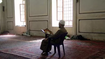 L'image montre un homme assis sur une chaise dans une mosquée. Il est vêtu d'une longue robe et porte un chapeau traditionnel. Devant lui, il lit un livre ouvert, probablement un exemplaire du Coran. La mosquée a un intérieur simple, avec des murs en marbre et des fenêtres laissant passer la lumière naturelle. Le sol est recouvert de tapis, et on peut voir d'autres personnes allongées au fond de la salle.