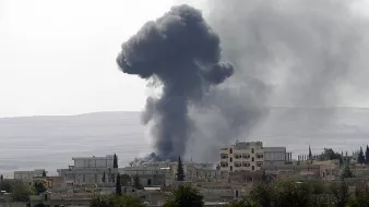 L'image montre un paysage urbain avec un nuage de fumée noir s'élevant dans le ciel, probablement à la suite d'une explosion ou d'un incendie. Les bâtiments aux alentours semblent endommagés, et la végétation est rare. Le fond présente des collines dégradées, ce qui suggère un environnement difficile. L'atmosphère générale évoque une situation de conflit ou de crise.