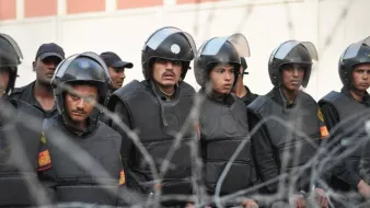 L'image montre un groupe de policiers en tenue anti-émeute, portant des casques et des gilets pare-balles. Ils semblent être en formation et regardent attentivement dans une direction, manifestant une posture de vigilance. En arrière-plan, on peut voir des barbelés, ce qui indique un contexte de sécurité renforcée ou de tensions. Les soldats portent des uniformes sombres et sont alignés, ajoutant à l'impression de discipline et de préparation.