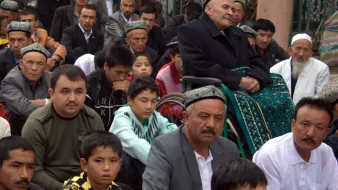 La imagen muestra a un grupo de personas sentadas en un ambiente al aire libre, posiblemente durante un evento comunitario o religioso. Se pueden ver hombres y niños, algunos de ellos vestidos con ropa tradicional. En el centro de la imagen, hay un hombre mayor sentado en una silla de ruedas, mirando hacia el frente. Los rostros muestran atención y respeto, reflejando la seriedad del momento. La escena parece estar en un lugar público, rodeado de otros espectadores.