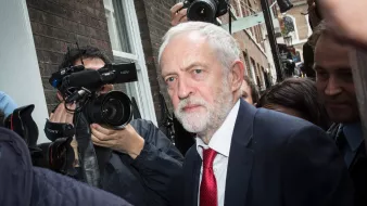 The image shows a man with gray hair and a beard, dressed in a dark suit and a red tie, walking through a crowd. He appears to be in a busy outdoor setting, surrounded by photographers and cameras capturing the moment. The buildings in the background suggest an urban environment. The overall atmosphere seems to convey a sense of public attention and media interest.