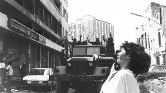 L'image montre une scène urbaine où une femme tient un appareil photo, prenant probablement des clichés d'une situation troublée. En arrière-plan, on peut voir un véhicule militaire sur une route, avec plusieurs personnes en uniforme visible sur le camion. Les bâtiments autour semblent en mauvais état, avec des stores métalliques fermés. L'atmosphère dégage une impression de tension ou de conflit. La femme, avec des cheveux bouclés, semble concentrée sur sa prise de vue, capturant un moment significatif dans un contexte difficile.