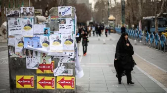 L'image montre une rue animée où une femme portant un hijab noir marche sur le trottoir. À côté d'elle, un mur est recouvert d'affiches et de banderoles, certaines de couleur vive, d'autres jaunies et déchirées. Les affiches semblent inclure des images de personnes et des slogans en écriture arabe. En arrière-plan, on peut distinguer des bus et des passants, créant une atmosphère urbaine. Le sol est en pavé, et les arbres dépouillés suggèrent une saison froide.