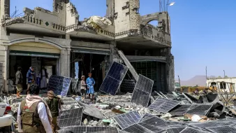 صورة لمكان مدمر، مباني مهدمة وألواح شمسية متكسرة، وأشخاص يتجمعون في الموقع.