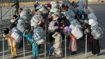 L'image montre un groupe de personnes, principalement des femmes, qui portent de grands fardeaux enveloppés dans des filets ou des tissus. Elles se trouvent dans un environnement urbain, probablement près d'un point de passage ou d'une frontière. La plupart des femmes sont vêtues de vêtements traditionnels et portent des foulards. Elles semblent attendre ou se regrouper, tandis qu'en arrière-plan, on aperçoit un soldat ou un garde. Cette scène évoque des thèmes de travail, de migration ou de transport de marchandises.