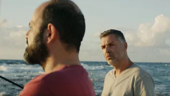 L'image montre deux hommes se tenant près de la mer. L'un d'eux a une barbe et porte un t-shirt rouge, tandis que l'autre a les cheveux grisonnants et un t-shirt clair. L'arrière-plan présente un océan calme sous un ciel bleu parsemé de nuages. L'atmosphère semble sereine, mais les expressions des hommes suggèrent une conversation intense ou réfléchie.