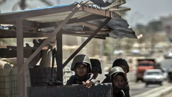 La imagen muestra a un grupo de soldados en un vehículo militar o en un puesto de control. Están vestidos con uniformes de combate y cascos, y algunos de ellos parecen estar armados. Al fondo, se pueden ver árboles y edificaciones, así como un cielo despejado. También hay un pequeño trozo de una bandera ondeando. La escena sugiere un ambiente de vigilancia o seguridad en una zona urbana.