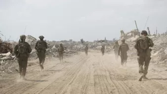 Soldiers in military gear walk through a dusty, desolate landscape with debris around them.