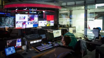 L'image montre une salle de contrôle de télévision ou de production, probablement un centre de diffusion. On y voit plusieurs écrans affichant des images en direct, des graphiques et des informations. Des personnes sont assises devant des consoles de contrôle, concentrées sur leur travail, et semblent superviser la diffusion. L'éclairage est tamisé, et on peut apercevoir une ambiance technologique avec des équipements modernes. Les murs en verre permettent d'apercevoir à l'extérieur, donnant une sensation d'ouverture.