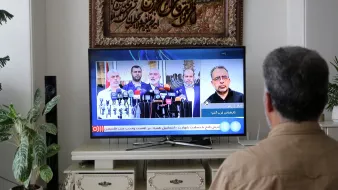 The image shows a person sitting in front of a television that displays a news broadcast. On the screen, several individuals are seen at a podium giving a press conference or statement, likely related to a political or social topic. The person is focused on the TV, and there is a decorative piece or artwork visible in the background, along with a plant near the television. The ambiance suggests a domestic setting where news is being followed closely.