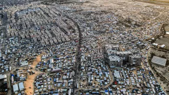 L'image montre une vue aérienne d'une vaste zone urbaine très densément peuplée, probablement une ville en développement ou un bidonville. On peut distinguer des habitations de différentes tailles et styles, de nombreuses structures temporaires, ainsi que des routes et des chemins qui traversent l'ensemble de la zone. La scène est marquée par un mélange de constructions durables et de tentes, suggérant une forte densité de population ainsi que des conditions de vie variées. En arrière-plan, on aperçoit des terrains plus dégagés, peut-être des zones agricoles ou industrielles.