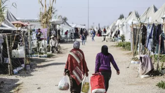 The image depicts two women walking down a dirt path in a makeshift camp. One woman carries bags of supplies, while the other pushes a stroller. On either side of the path, there are tents and structures that suggest a temporary living situation. The environment appears to be bustling with activity, as other individuals can be seen in the background, some seated or engaging in conversation. The scene conveys a sense of community within the challenges of a refugee or displaced persons camp.