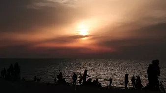 The image depicts a beach scene during sunset. In the foreground, there are silhouettes of people walking along the shore, some sitting or standing by the water. The sky is filled with soft hues of orange and pink, blending into darker clouds as the sun sets on the horizon. The water reflects the colors of the sky, creating a tranquil and serene atmosphere. Overall, the scene conveys a sense of calm and natural beauty as day transitions into night.