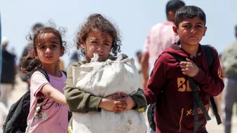 L'immagine mostra un gruppo di bambini che camminano, visibilmente in una situazione di difficoltà. Due bambine, una con i capelli ricci e l'altra con un'espressione preoccupata, accompagnano un ragazzo che tiene una grande borsa. Sembra che si trovino in un contesto di emergenza, con altre persone sullo sfondo. L'atmosfera è seriosa e trasmette una sensazione di vulnerabilità e speranza.