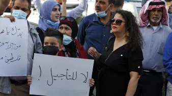 L'image montre un groupe de personnes rassemblées pour une manifestation. Parmi elles, il y a des adultes et des enfants, certains portant des masques. Une femme en noir se tient à droite, tandis qu'un jeune garçon au centre tient une pancarte avec un message en arabe. Les manifestants semblent exprimer des préoccupations ou des revendications, chacun affichant une expression déterminée. L'ambiance générale est celle d'une protestation pacifique.