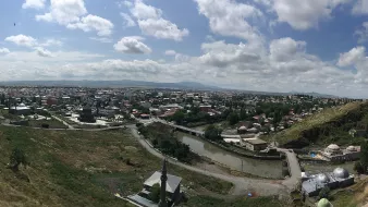 L'image représente une vue panoramique d'une ville avec un paysage verdoyant. On peut voir des toits de maisons, des bâtiments et des infrastructures qui s'étendent à travers la vallée. Le ciel est partiellement nuageux, laissant passer la lumière du soleil. Au premier plan, il y a des collines et des routes qui serpentent à travers le paysage. L'ensemble dégage une atmosphère paisible et naturelle, avec une harmonie entre l'urbanité et la nature environnante.