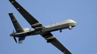 L'image montre un drone de type militaire, en vol. Il a une conception aérodynamique avec des ailes étendues et une queue distinctive. La surface du drone est peinte en gris, ce qui le rend moins visible en altitude. On peut également observer des équipements sous ses ailes, probablement destinés à la surveillance ou à des missions de reconnaissance. Le ciel en arrière-plan est dégagé, mettant en valeur le drone en mouvement.