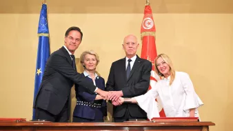 L'image montre quatre personnes se serrant la main autour d'une table. À gauche, un homme en costume sombre, probablement un représentant d'un pays européen. À sa droite, une femme avec des cheveux blonds peignés, semblant être une dirigeante politique. Au centre, un homme en costume sombre, certainement le président d'un pays, et à sa droite, une femme vêtue de blanc, probablement une autre dirigeante. En arrière-plan, on peut voir des drapeaux de l'Union européenne et de la Tunisie, ce qui suggère une rencontre diplomatique.