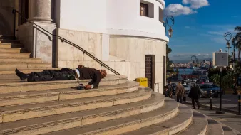 L'image montre un homme allongé sur les marches d'un bâtiment au style architectural classique, probablement en train de dormir ou en état de détresse. Le bâtiment est bien éclairé et ses murs sont de couleur claire, avec des colonnes visibles. En arrière-plan, on peut apercevoir une ville animée, avec des gens qui marchent et un port. Le ciel est bleu avec quelques nuages, ce qui donne une ambiance contrastée à la scène.