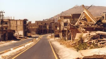 L'image montre une scène de destruction et de désolation. On peut voir une route déserte, bordée de bâtiments en ruines et de débris. Les structures semblent fortement endommagées, avec des murs effondrés et des fondations exposées. À l'arrière-plan, une colline se dresse, mais l'accent est principalement mis sur la destruction des habitations. L'atmosphère est empreinte de tristesse et d'abandon, suggérant que cet endroit a subi des conflits ou des catastrophes.