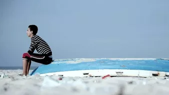 L'image montre un jeune garçon assis sur une coque de bateau échouée sur une plage. Il porte un t-shirt à rayures et un pantalon rouge. L'environnement est clair, avec un ciel bleu pâle et une plage de sable. Le garçon semble pensif, tourné vers l'horizon, tandis que la coque du bateau, abandonnée et usée, ajoute un élément de mélancolie à la scène.