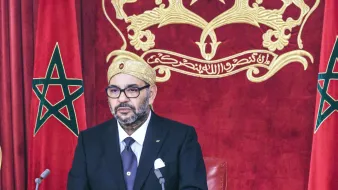 L'image montre un homme assis à un bureau, habillé élégamment en costume noir avec une cravate. Il porte un turban ou un couvre-chef traditionnel. En arrière-plan, on peut voir deux drapeaux marocains ornés d'une étoile à cinq branches. Le décor est riche, avec un fond rouge et une représentation du blason du Maroc. L'homme semble concentré et prêt à s'exprimer.