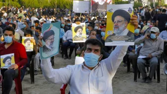 The image shows a large crowd of people seated outdoors, wearing masks. Many attendees are holding posters or pictures of a man, who appears to be a political figure, indicating support for him. The setting looks like a public gathering or rally, with trees in the background and banners visible. The mood appears to be one of engagement and solidarity among the participants.