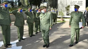 L'image montre un groupe de militaires en uniforme, saluant un supérieur. Tous portent des masques et des casquettes, ce qui indique qu'ils respectent des protocoles de santé. L'environnement semble être un site officiel ou militaire, avec des bâtiments en arrière-plan. Les militaires adoptent une posture de respect, ce qui suggère une cérémonie ou un événement formel.