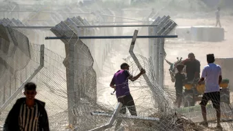 تصویر نشاندهنده مردانی است که در حال عبور از حصاری فلزی و برقراری یا تخریب موانع هستند. گرد و غبار در هواست.