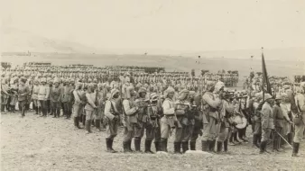 The image depicts a large group of soldiers lined up in a formation during a military event, likely a parade or review. The soldiers are dressed in uniforms typical of early 20th-century military attire, complete with helmets and boots. Some are holding rifles, and there are banners or flags present. In the background, more soldiers can be seen in organized rows, suggesting a significant military presence. The setting appears to be an open field, with hills or mountains visible in the distance, indicating a potentially historic or ceremonial occasion. The image has a monochromatic tone, typical of photographs from that era.