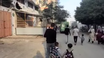 A man walks with two children on a dusty street, while others stroll nearby. Buildings line the path.