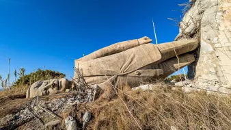 L'image montre une statue gigantesque de la main et du visage d'un personnage allongé sur le sol, entourée de verdure et sous un ciel bleu. La statue semble partiellement détruite ou en ruine.