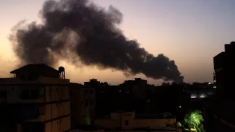 La imagen muestra un paisaje urbano al atardecer, con un cielo que presenta tonos oscuros. En el fondo, se puede ver una densa columna de humo negro que se eleva, sugiriendo la presencia de un incendio o alguna actividad industrial. A la izquierda, hay edificios que se recortan contra el cielo, y a la derecha, unas palmeras iluminadas por luces artificiales. La atmósfera parece tensa debido al humo y el entorno oscuro.