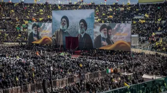 L'image montre une grande foule rassemblée dans un stade, avec des étendues de drapeaux de différentes couleurs. Au centre, trois grandes affiches de figures importantes sont visibles, probablement des leaders religieux ou politiques. L'atmosphère semble festive et engagée, avec des personnes portant des chapeaux ou des foulards spécifiques, suggérant une démonstration de soutien ou de solidarité. Des bannières et des drapeaux supplémentaires peuvent également être aperçus, accentuant l'aspect coloré et vibrant de l'événement.