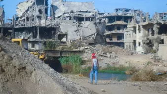 L'image montre un paysage de destruction, avec des bâtiments gravement endommagés, laissant apparaître des structures en béton et des débris éparpillés. Au premier plan, on aperçoit une personne qui semble marcher près d'une étendue d'eau, portant un chapeau rouge. L'environnement est déserté, avec une végétation qui essaie de reprendre ses droits parmi les ruines. Le ciel est bleu, mais le contraste entre la couleur du ciel et la destruction environnante crée une atmosphère de désolation.