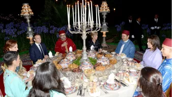 L'image montre une scène de dîner élégant, probablement dans un cadre extérieur. Une grande table est dressée avec une variété de plats et de délicieuses spécialités culinaires. De nombreuses bougies sont allumées, créant une ambiance chaleureuse et festive. Des convives, habillés en vêtements traditionnels, sont assis autour de la table, engageant des discussions animées. Au fond, on peut apercevoir des éléments décoratifs et des personnes en costume qui ajoutent à l'atmosphère formelle de l'événement.