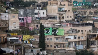 L'image montre un paysage urbain d'une ville, où plusieurs bâtiments sont alignés sur une colline. À travers ces constructions, on peut voir de grandes peintures murales représentant des yeux, aux couleurs vives, qui ajoutent une dimension artistique à cet environnement. Les bâtiments varient en couleurs et en textures, allant du beige au rose, et certains semblent en cours de rénovation. L'atmosphère générale évoque un mélange de quotidien et de créativité, avec une forte présence d'art urbain.