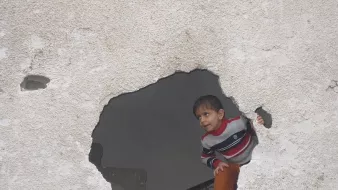 L'image montre un enfant qui regarde à travers un trou dans un mur. Le mur est en mauvais état, avec une surface rugueuse et des fissures visibles. L'enfant porte un pull rayé et semble curieux, se penchant légèrement pour observer ce qui se trouve de l'autre côté du mur. La scène évoque une atmosphère d'exploration et d'intrigue.