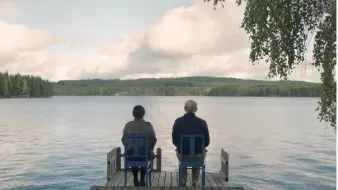 L'image montre deux personnes assises sur des chaises en bois sur une jetée qui s'avance dans un lac paisible. Ils regardent vers l'horizon, avec des arbres verdoyants en arrière-plan et un ciel nuageux au-dessus. L'atmosphère est calme et sereine, évoquant un moment de contemplation ou de discussion tranquille.