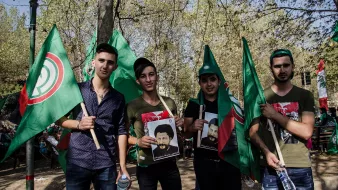 L'image montre un groupe de quatre jeunes hommes rassemblés lors d'un événement, probablement une manifestation ou une célébration. Ils tiennent des drapeaux verts, qui semblent avoir un symbole spécifique. Chacun d'eux porte des affiches représentant des figures, et ils affichent des expressions de fierté ou de détermination. L'arrière-plan est verdoyant, évoquant un cadre festif en plein air.