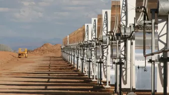 L'image montre une série de panneaux solaires en concentrateurs, alignés sur un terrain désertique. Ces structures lumineuses captent la lumière du soleil pour générer de l'énergie. En arrière-plan, on peut voir un ciel partiellement nuageux avec des montagnes lointaines. L'environnement semble aride, avec des collines de terre et un véhicule de construction visible à distance. Les ombres des panneaux s'étendent sur le sol, soulignant leur orientation vers le soleil.