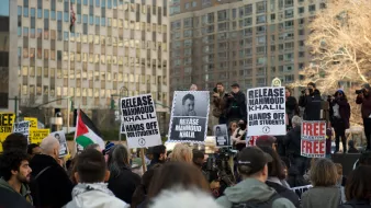 Des manifestants se rassemblent dans une ville, tenant des panneaux avec des slogans, notamment pour la libération de Mahmoud Khalil et des revendications pour les droits des étudiants. En arrière-plan, on peut voir des bâtiments et des personnes utilisant des caméras. L'ambiance est déterminée et engagée.