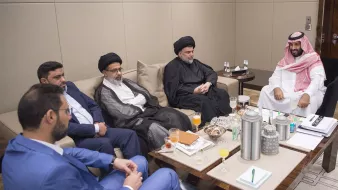 L'image montre un groupe de personnes assises autour d'une table dans une pièce moderne et élégante. Ils semblent engagés dans une discussion sérieuse. Sur la table, il y a des boissons et des collations. Les participants portent des vêtements formels, certains avec des vêtements traditionnels, ce qui souligne le contexte culturel de la réunion. L'ambiance est professionnelle et conviviale.